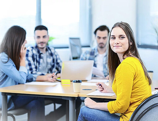 Junge Frau, die mit mehreren Personen an einem Tisch sitzt und in die Kamera lächelt